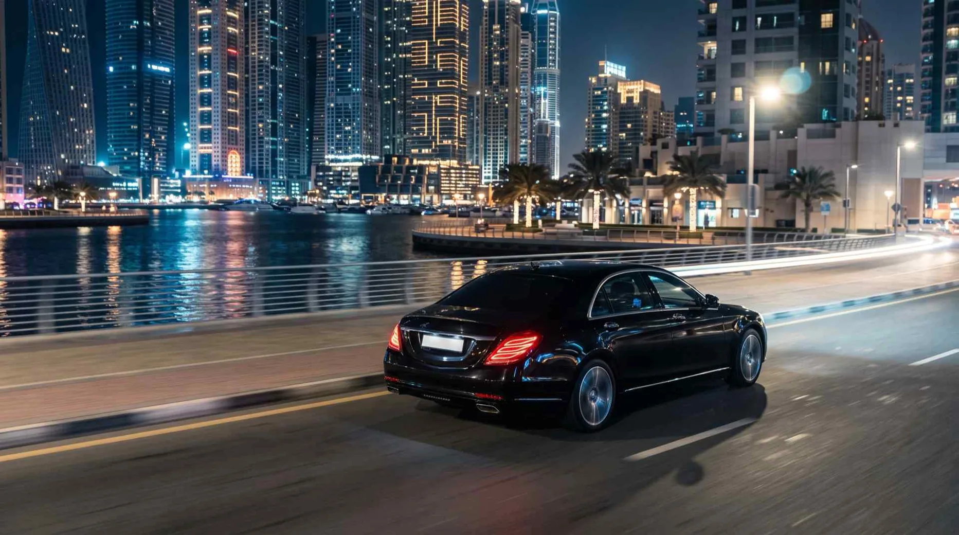 A black sedan driving along a waterfront road at night, lit towers behind